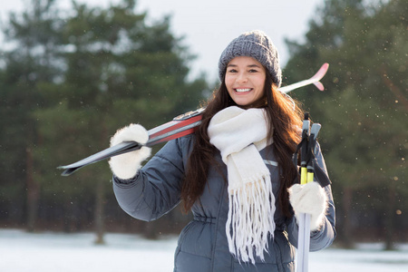 微笑的女孩在灰色针织豆帽, 白色围巾和蓬松的手套举行滑雪板和棍子