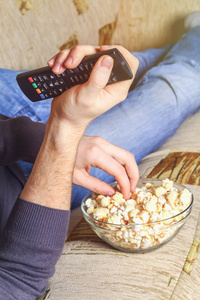 一个男人拿着一碗爆米花, 手里拿着遥控器看着沙发上的电视。