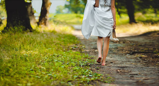穿着白色连衣裙的新娘赤脚沿着公园的小路走着女孩从高跟鞋休息照片