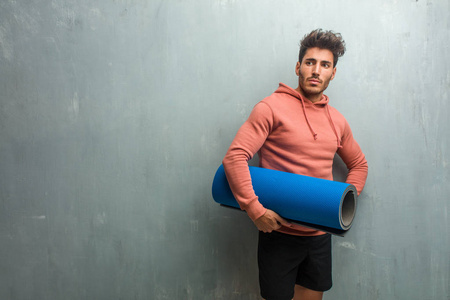 年轻的健身男子对一个垃圾墙交叉他的胳膊, 拿着一个蓝色的垫子练习瑜伽