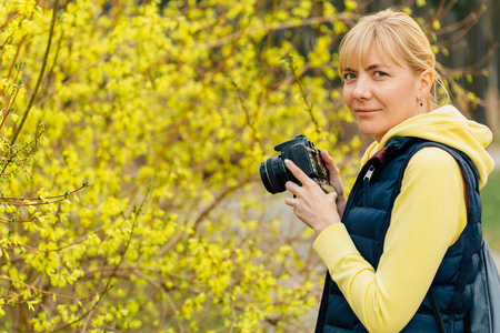 女孩与专业相机在手站立附近连翘灌木。公园女摄影师特写
