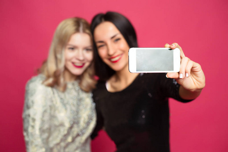 两个开朗和美丽的微笑的女朋友拍自己在电话或做一个自拍在粉红色的背景