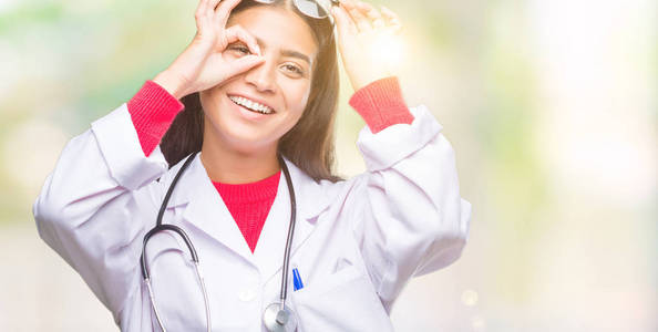 年轻的阿拉伯医生妇女在孤立的背景下做 ok 手势用手微笑, 眼睛看通过手指与愉快的面孔