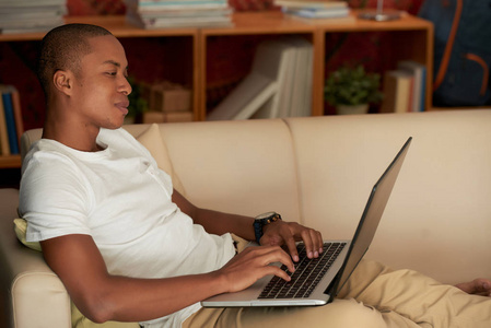 年轻男子坐在沙发上, 在他的客厅和工作的笔记本电脑