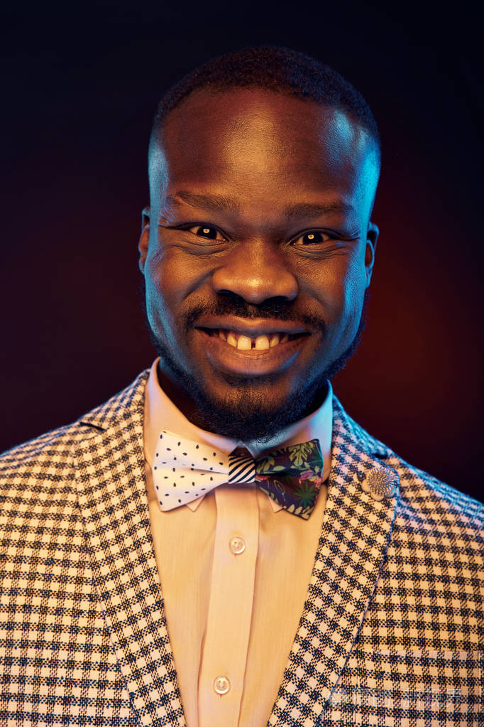 工作室肖像微笑英俊的黑人与滑稽的面孔在白色衬衣和格子夹克与花弓