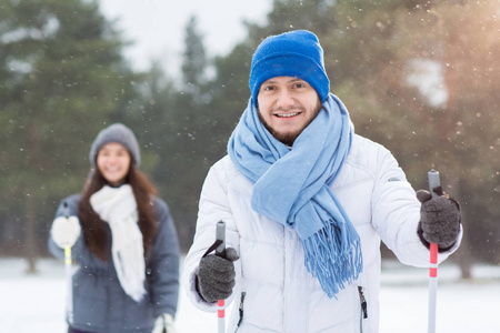 微笑的年轻人在蓝色豆帽和围巾举行滑雪棍子和享受滑雪在雪