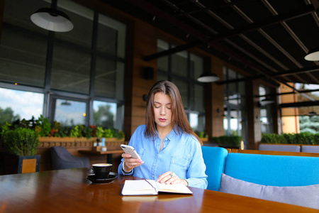 女孩保持智能手机, 阅读笔记本和休息在咖啡馆与咖啡