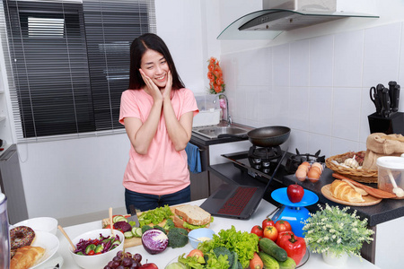 快乐的妇女烹饪和寻找与笔记本电脑在厨房里