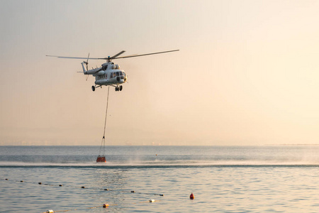 一架带红色篮子的直升飞机在海面上降落, 在黎明的橙色天空和远处的城市剪影上舀水。