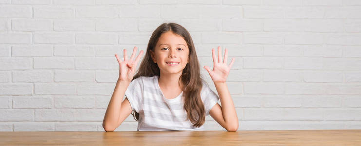 年轻的西班牙裔孩子坐在家里的桌子上显示和指着手指九号同时微笑自信和快乐