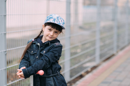 可爱的小女孩在牛仔裤羊毛衫和踏板车的背景上的围栏