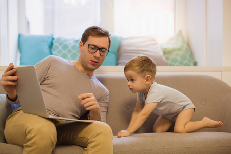 积极的年轻父亲坐在沙发上, 在房间附近, 他可爱的小儿子, 他们看笔记本电脑