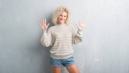 年轻的金发女人与卷曲的头发在垃圾灰色背景显示, 并指出手指十, 而微笑着自信和快乐