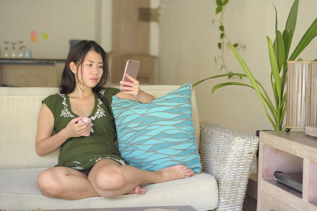 年轻美丽和轻松的亚洲华人妇女躺在家里的客厅沙发沙发上使用互联网上的手机快乐和舒适