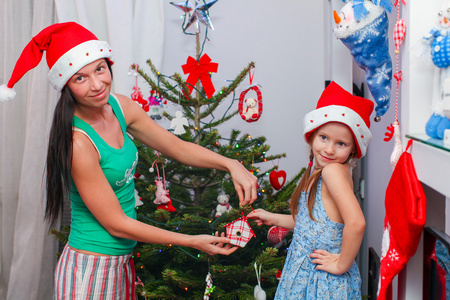年轻的母亲和她可爱的小女孩在圣诞老人帽子装饰他们的圣诞树