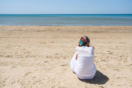 孤独的黑发女人, 戴着围巾坐在海滩上, 看着大海。后视图