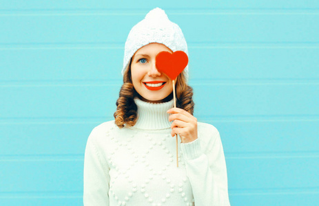 肖像快乐微笑年轻女子抱棒糖棒心形在针织白帽, 毛衣在蓝色背景下