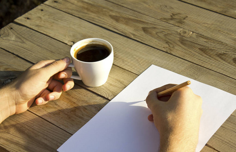 男性手拿着咖啡杯, 在白纸上写字, 木桌