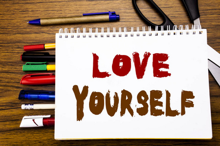 字, 写爱自己。商业概念为积极的口号为您写在笔记本上, 木制背景与办公设备如钢笔剪刀彩色标记