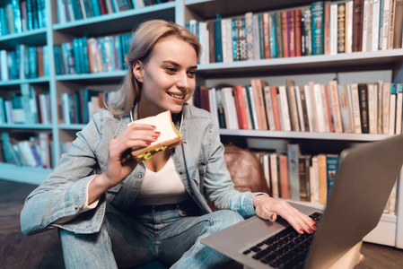 白色女孩坐在图书馆的书架附近, 吃三明治和使用笔记本电脑