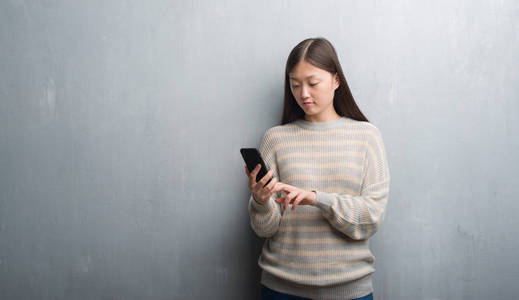 灰色墙上的年轻中国妇女看着智能手机与自信的表情, 认真思考