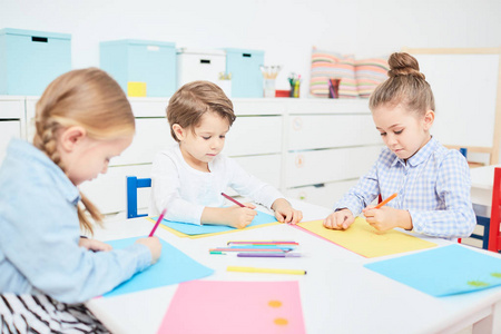 可爱的年轻孩子坐在桌子上, 在幼儿园画画与蜡笔