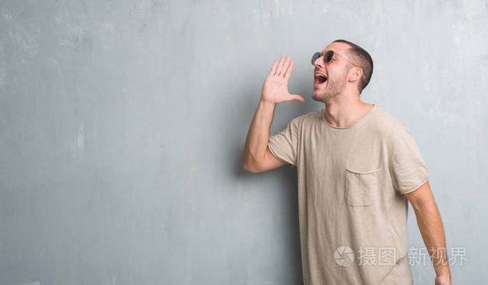 年轻的白种人在灰色的垃圾墙上戴着太阳镜大喊, 大声尖叫到一边用手在