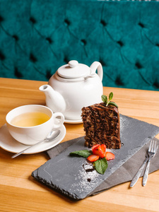 巧克力斑马蛋糕在石板上配有草莓切片, 薄荷叶, 糖粉, 杯茶, 茶壶, 叉子和勺子放在木桌上。