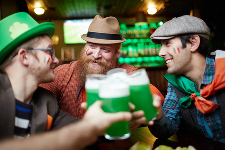 快乐的年轻同伴坐在酒吧里, 用绿色的啤酒在酒杯里举杯庆祝。