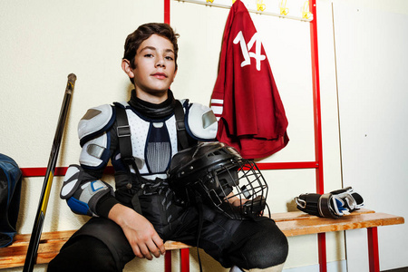 在男子冰球更衣室里坐在长凳上的保护装备少年男孩特写肖像