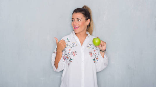 年轻的成年妇女在灰色的难看的东西墙壁吃新鲜的绿色苹果指向和显示与拇指高兴脸微笑