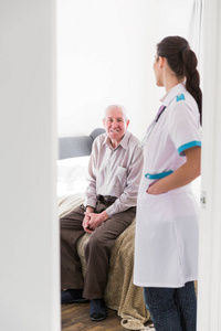 坐在家里的一张床上的老微笑的男人和年轻的护士站在一个房间附近的门