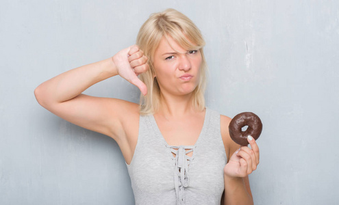 白人成年妇女在灰色的垃圾墙上吃巧克力甜甜圈与愤怒的脸, 消极的迹象显示不喜欢拇指下来, 拒绝概念