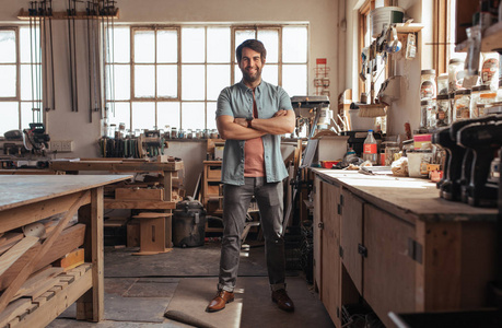 年轻的木工的肖像微笑着自信, 而站在他的手臂交叉工作台在他的木工车间充满工具
