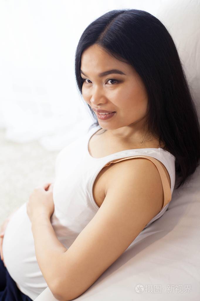 美丽的年轻的亚洲孕妇坐在窗口和触摸她的肚子怀孕健康母亲一个孕妇的