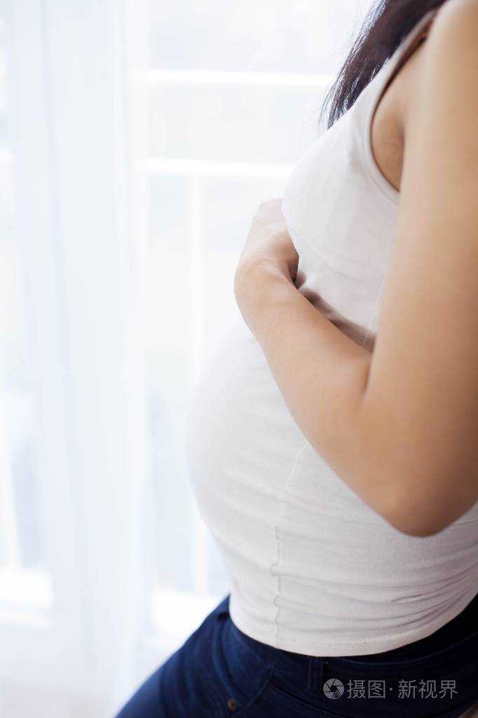 美丽的年轻亚洲孕妇站在窗口和触摸她的肚子.怀孕.健康.母亲.