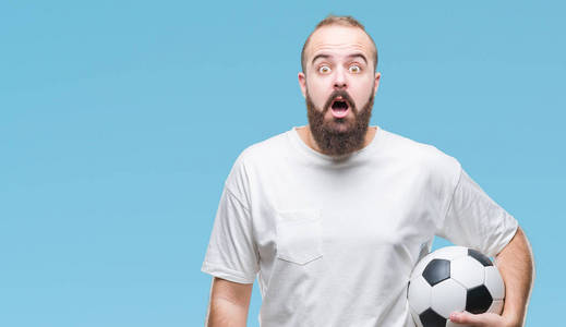 年轻的高加索嬉皮士男子拿着足球在孤立的背景吓得惊心动魄的一张惊讶的面孔, 害怕和兴奋与恐惧的表情