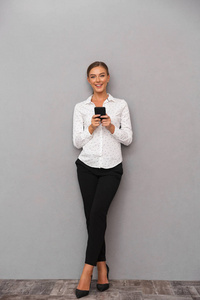 美丽的微笑的企业妇女的形象站在灰色墙背景上使用手机