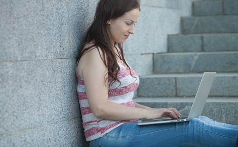 一个快乐的年轻女子坐在楼梯上的城市和户外使用便携式计算机的肖像