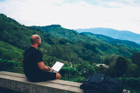 在山区夏季徒步旅行中坐在高山上的数字上网本中, 男性旅游者对绿色景观的欣赏性