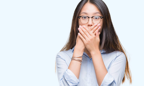 年轻的亚洲商业妇女戴着眼镜在孤立的背景震惊地覆盖口与手错误。秘密概念