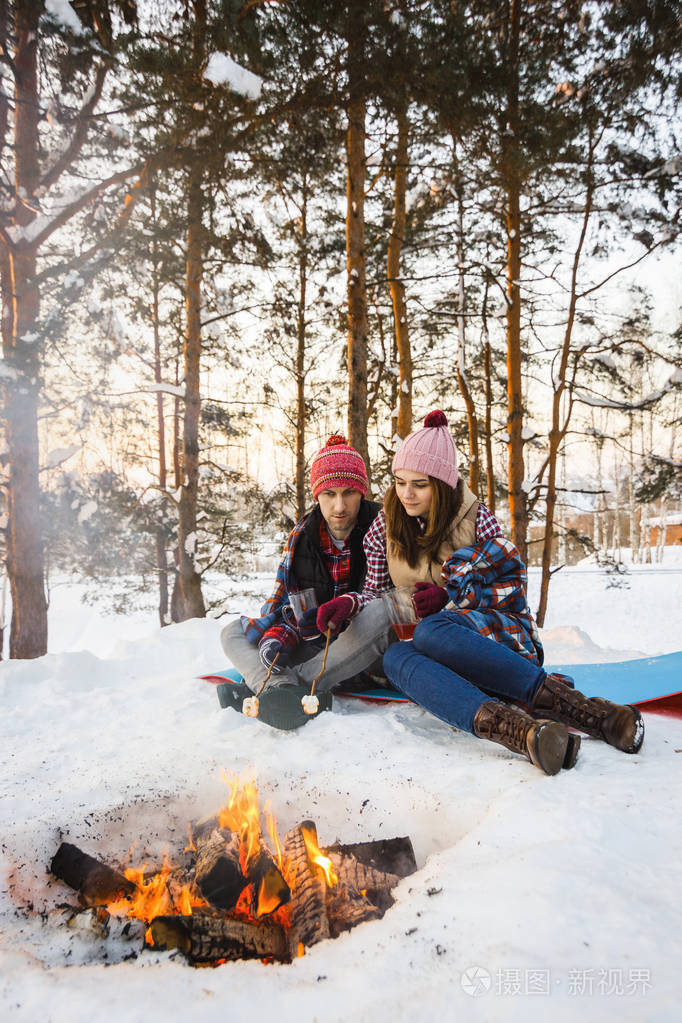年轻夫妇煎棉花糖在冬天的火在森林下的地毯
