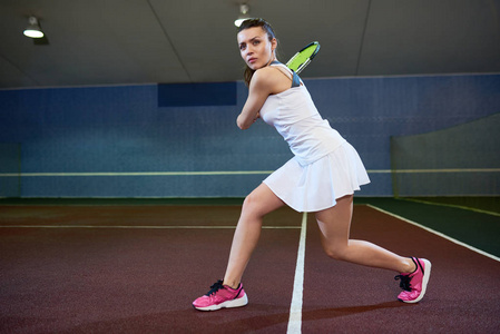 全长肖像自信的年轻女子在室内球场打网球, 准备打飞球