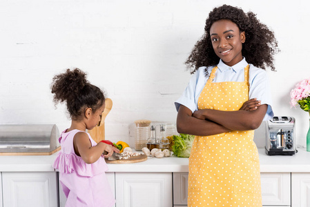 非洲裔美国人的母亲站在交叉的怀抱, 而女儿在厨房切菜