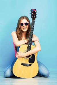 时尚音乐与年轻模型在太阳镜中抱着原声吉他, 坐在蓝色的地板上