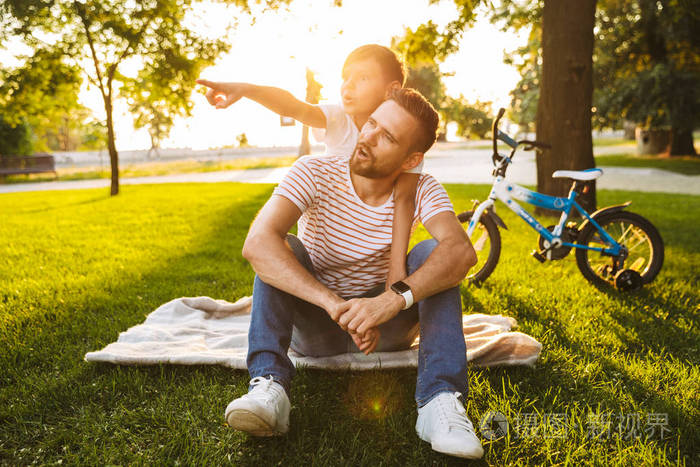 兴奋的父亲和他的儿子一起在绿色公园的乐趣, 坐在毯子上, 背着骑, poitning 离开