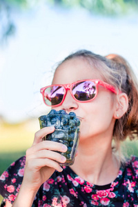 快乐的女孩享受新鲜的蓝莓户外。少女穿着华丽的红蓝上衣和红色的太阳镜。庭院在背景