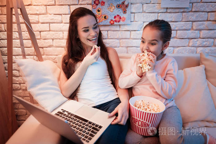 母亲和小女儿看电影在笔记本电脑和吃爆米花在晚上在家