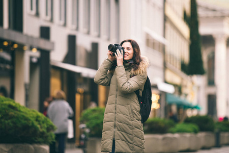 美丽的年轻旅游妇女站立在中央街道的背景在慕尼黑在德国在冬天。持有黑色大的专业相机, 拍照和微笑