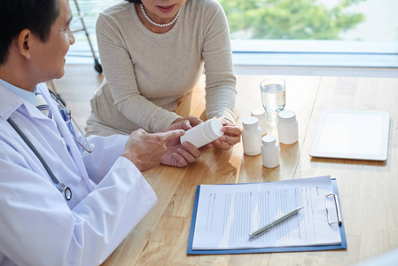 友好的亚洲心脏病专家坐在桌旁, 解释他的女性病人如何服药照片